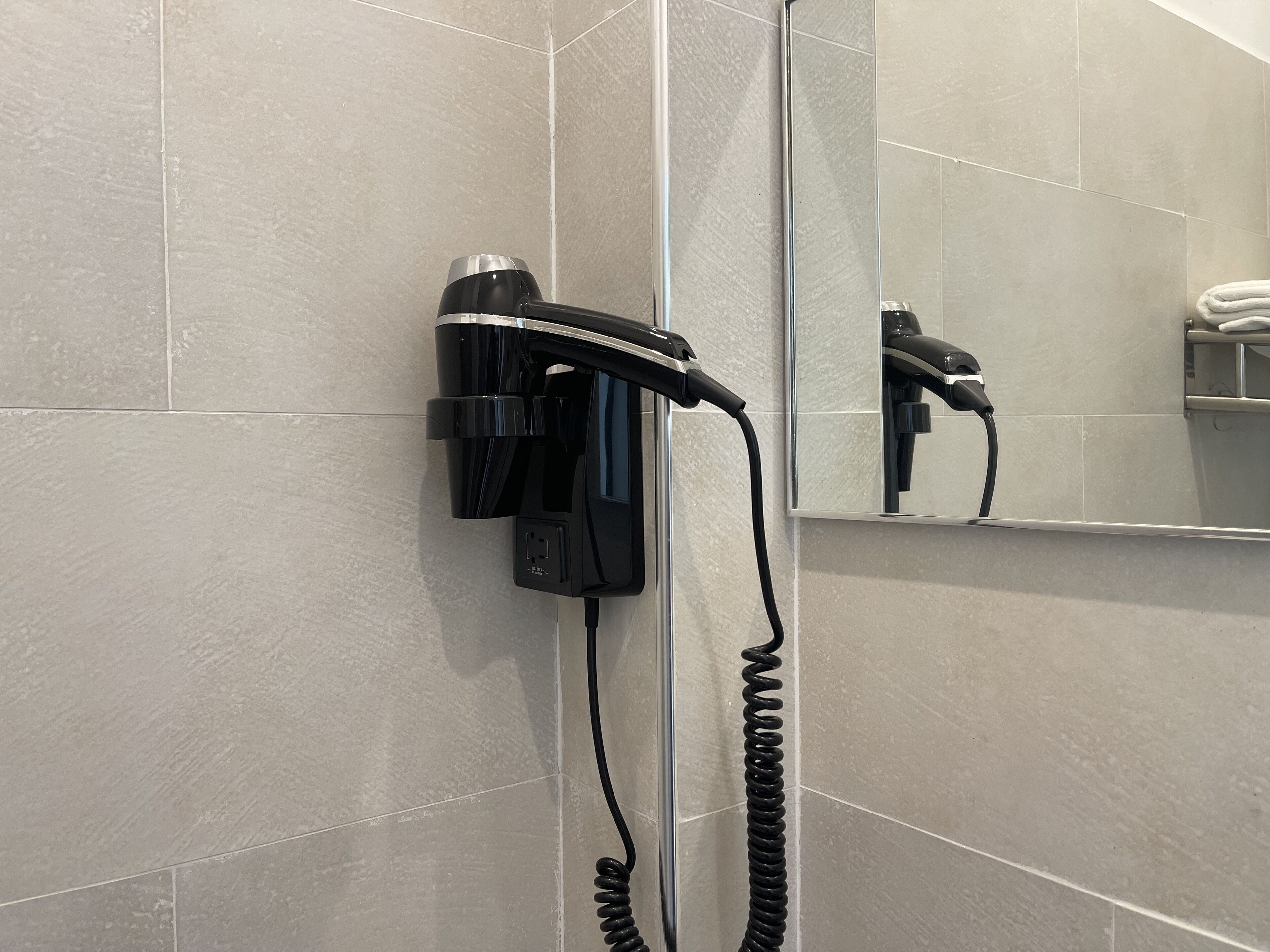 Un secador de pelo montado en la pared de un baño con paredes de azulejos y un espejo que refleja el secador.