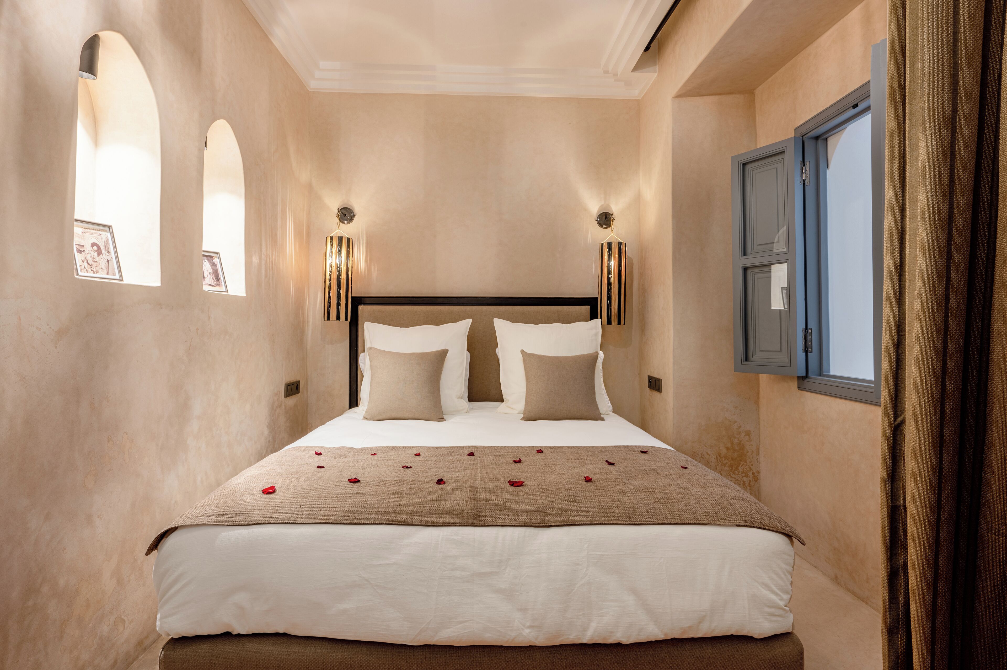 Ein ordentlich bezogenes Bett mit einer beigen Decke und Kissen, zwei Wandleuchten und ein Fenster mit Vorhängen.