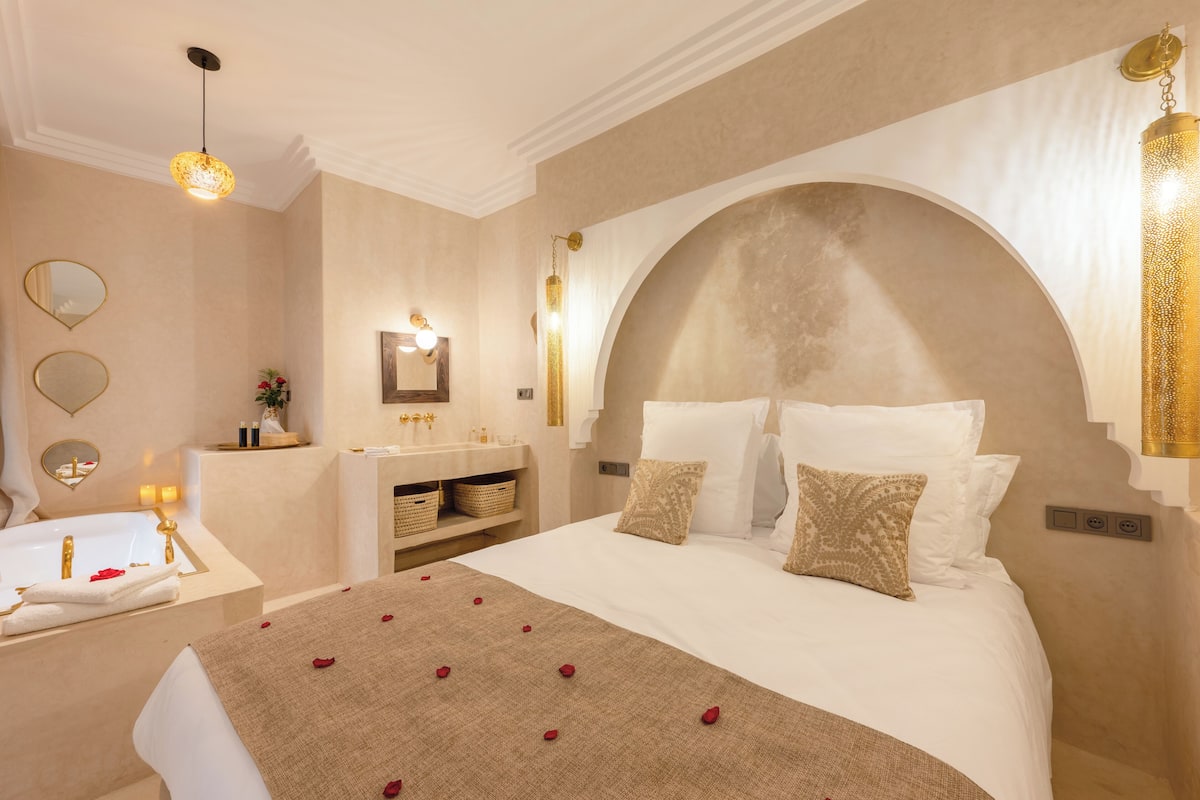 A hotel room with a large bed, a vanity with a mirror, a bathtub, and a large arched window.