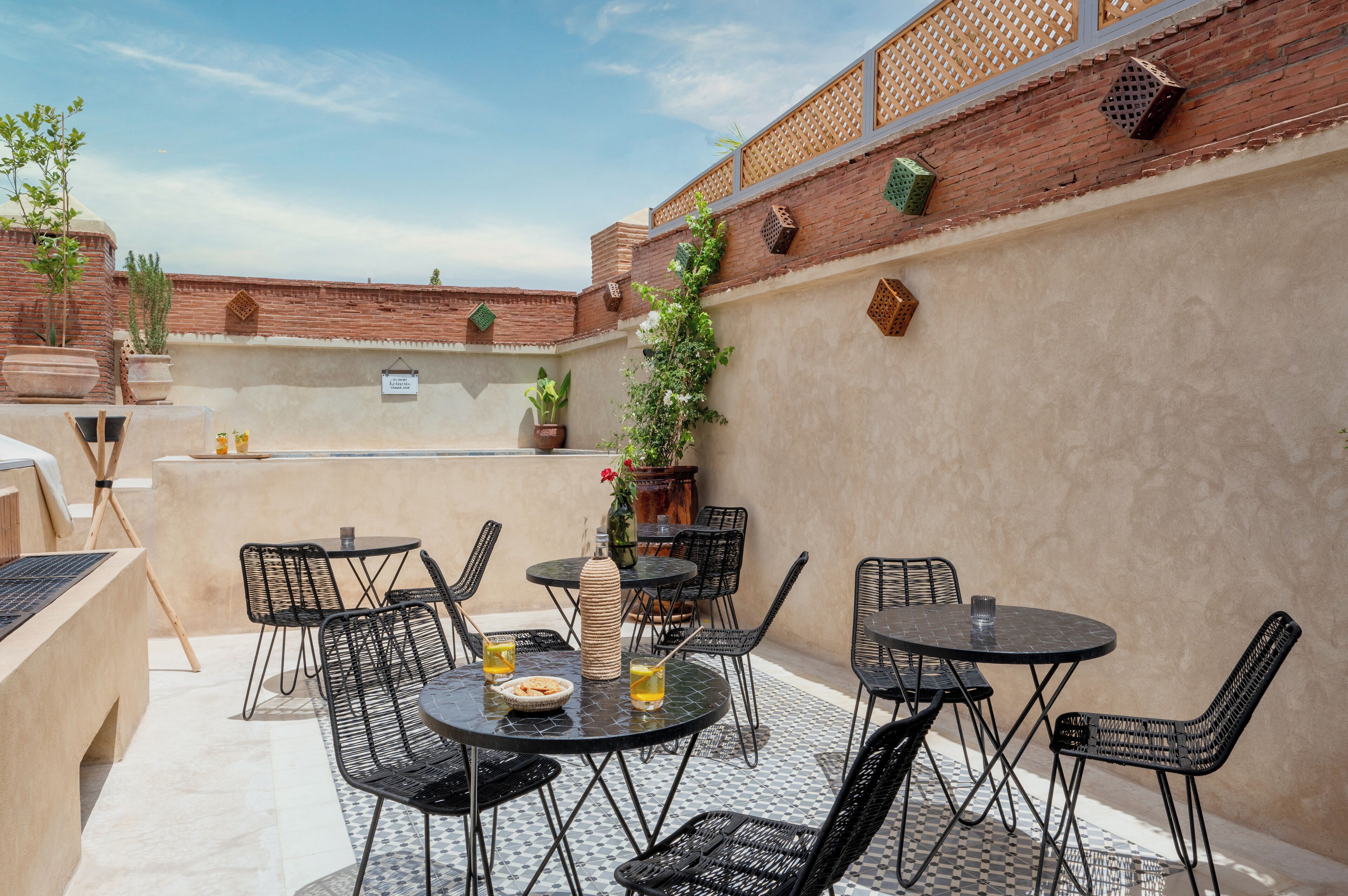 An outdoor patio with round tables and black metal chairs, surrounded by potted plants and a brick wall.