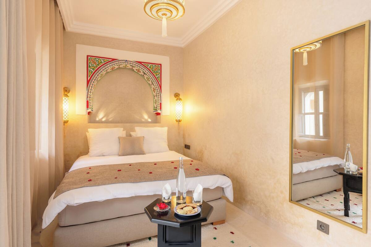 A hotel room with a large bed, a bedside table with a bottle and glasses, and a decorative arch above the bed.