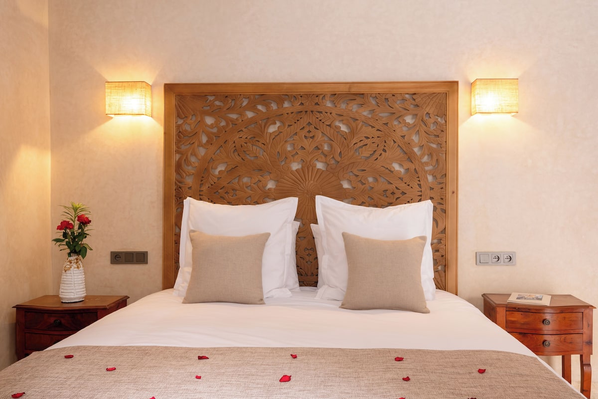 A neatly made bed with a decorative headboard, two bedside tables, and a vase with flowers.
