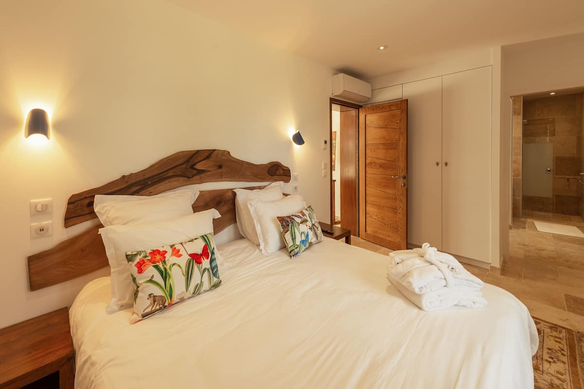 A neatly made bed with white linens and decorative pillows, a wooden headboard, and a wall-mounted light fixture.