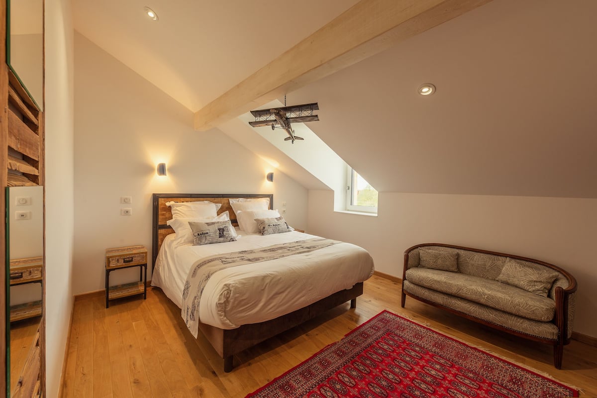 A bedroom with a wooden floor, a bed with white bedding, a red rug, a wooden bench, and a sloped ceiling with a hanging light fixture.