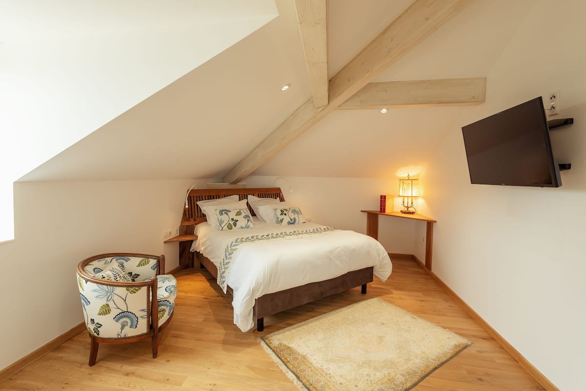 A bedroom with a sloped ceiling, a bed with white bedding, a wooden chair, a bedside table with a lamp, and a TV mounted on the wall.