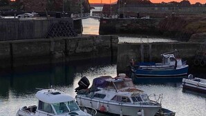 A harbor with boats docked, a bridge, and a rocky cliff in the background during sunset.