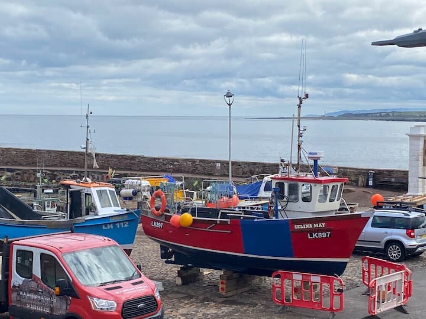 A harbor with boats docked, a red van, and a stone wall in the background.