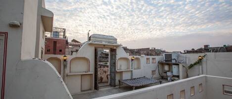 A rooftop terrace with a view of the cityscape, featuring a mix of traditional and modern architecture, including a distinctive white building with a flat roof and a small garden area.