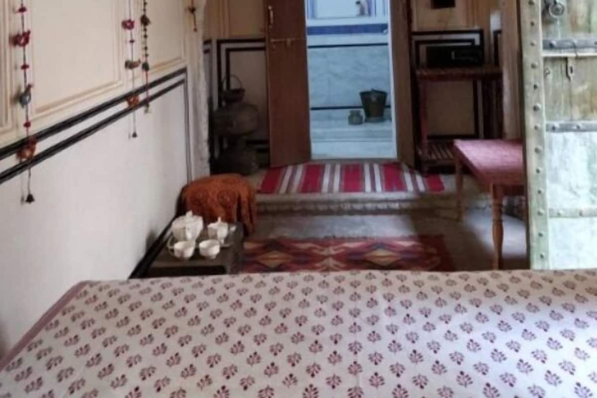 Una habitación con cama, ventilador de techo, una mesita con mantel rojo y blanco, y vistas a un balcón por un umbral abierto.
