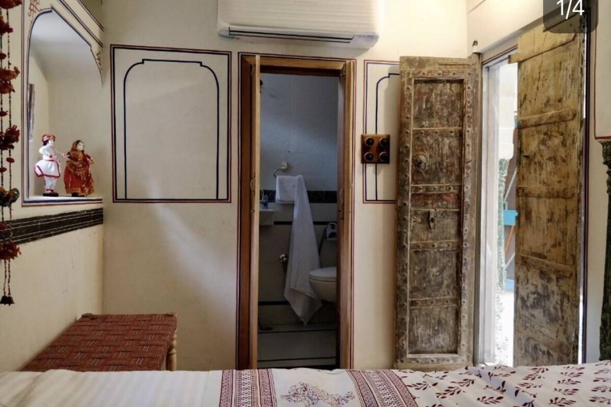 Un dormitorio con cama, una mesita, un armario y un baño a la vista a través de una puerta abierta.