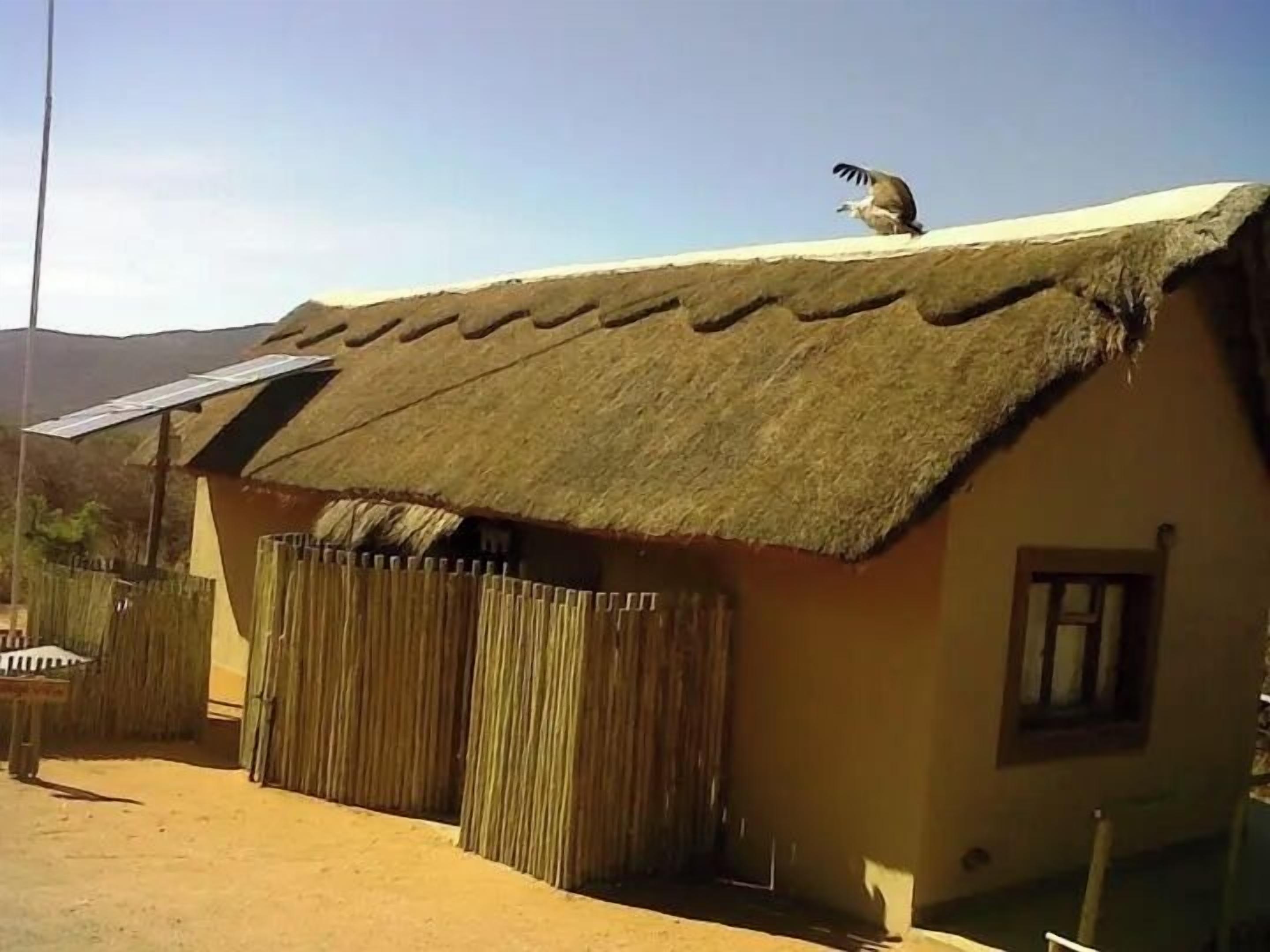 A thatched-roof house with a solar panel on the roof, a wooden fence, and a mountain in the background.