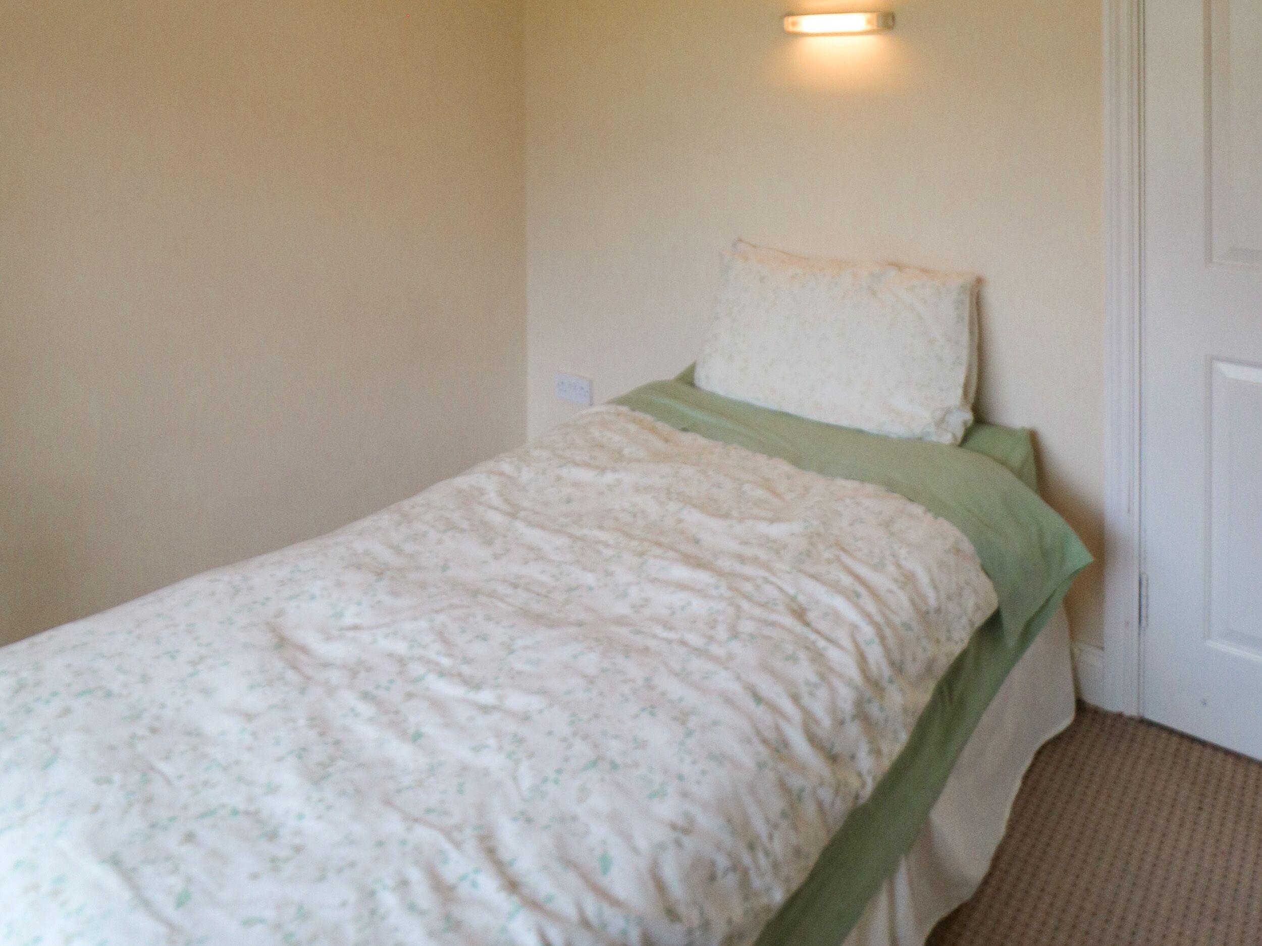 A single bed with a floral-patterned bedspread and a green pillow in a room with beige walls and a white door.