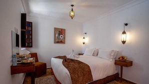 A hotel room with a large bed, a desk with a water bottle and a tray, a chair, two wall-mounted lights, and a painting on the wall. - Riad Dar Nejma & Spa (Marrakech)