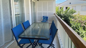 Un balcone con tavolo in vetro e sedie blu, ringhiera bianca e vista su spazi verdi e edifici.