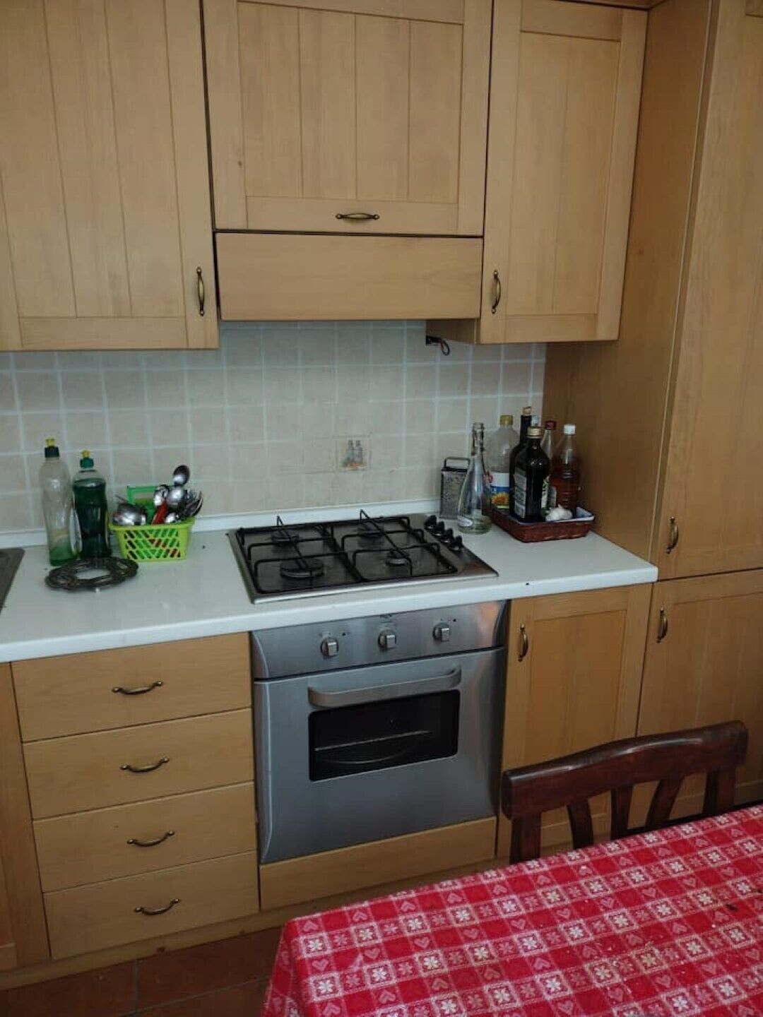 Une cuisine avec des meubles de rangement en bois, une cuisinière à gaz et un évier. On peut voir des bouteilles et un panier vert sur le plan de travail.