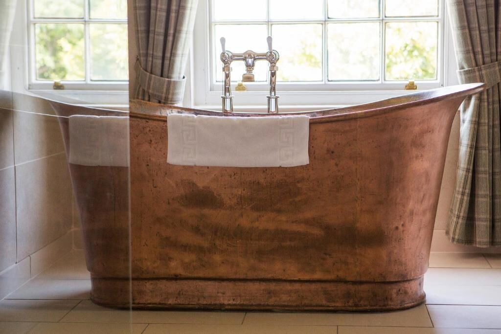 Una bañera de cobre antigua con una toalla blanca, cerca de una ventana con cortinas.