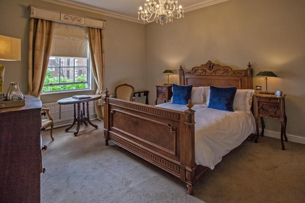 A classic bedroom with a wooden bed, two bedside tables, a chandelier, and a view of greenery outside.