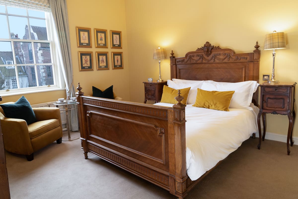 A bedroom with a wooden four-poster bed, a small round table, a chair, and a window with curtains.
