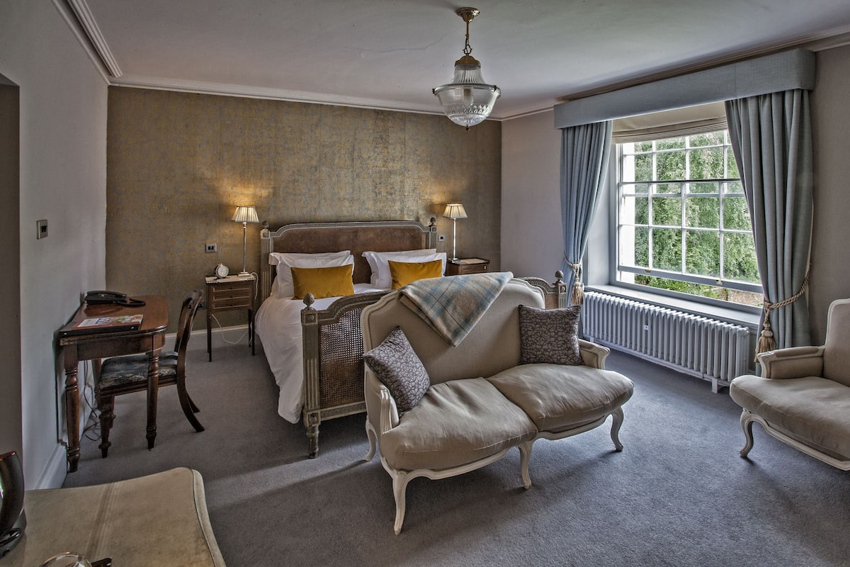 A classic hotel room with a large bed, two armchairs, a wooden nightstand, a desk with a lamp, and a view of greenery through the window.