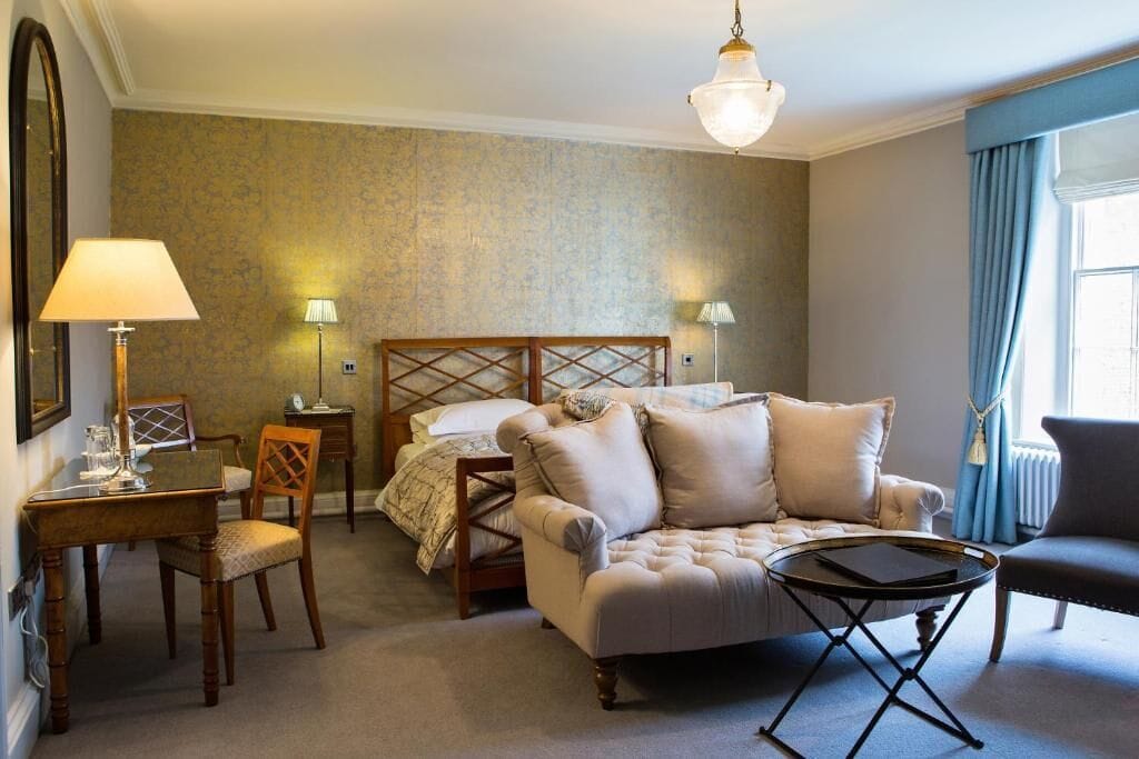 Et hotelværelse med seng, sofa, spisebord og stol. Der er lamper på hver side af sengen og et vindue med gardiner.
