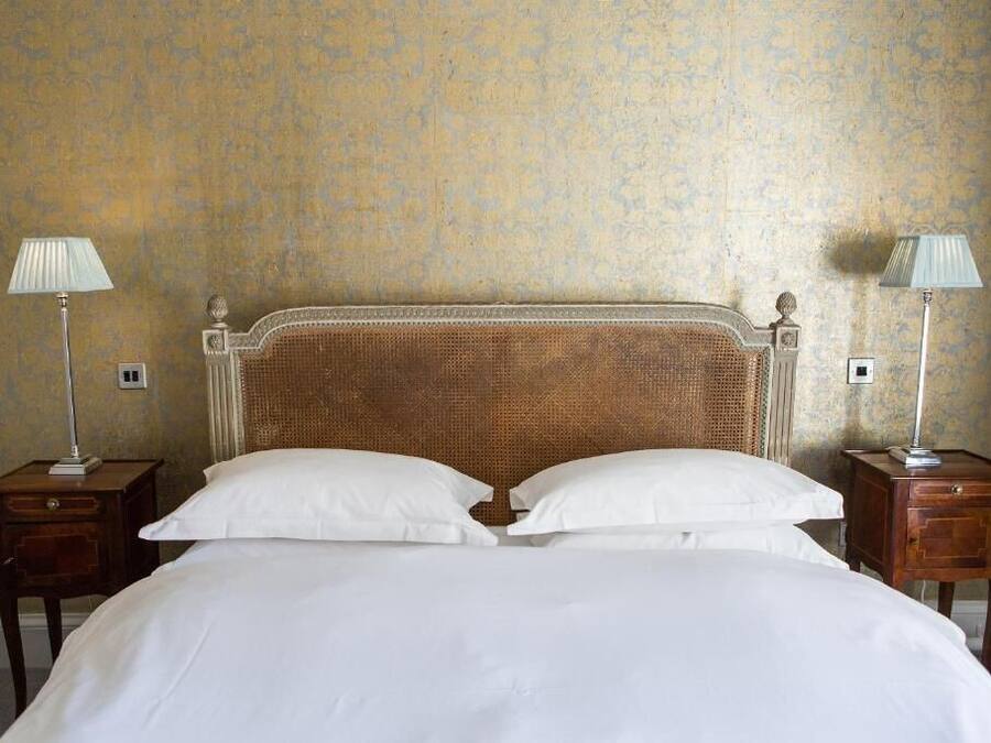 Una cama bien hecha con sábanas blancas, flanqueada por dos mesitas de noche con lámparas, frente a una pared con textura.