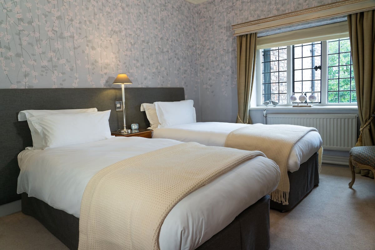 A hotel room with two beds, a window with curtains, a lamp, and a radiator.