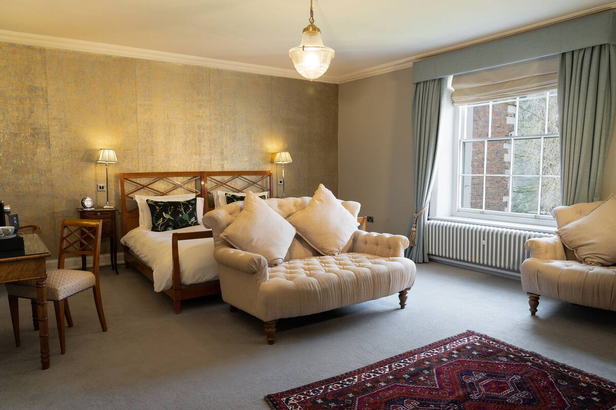 A bedroom with a large bed, two bedside tables, a wooden chair, a sofa, a patterned rug, and a window with curtains.
