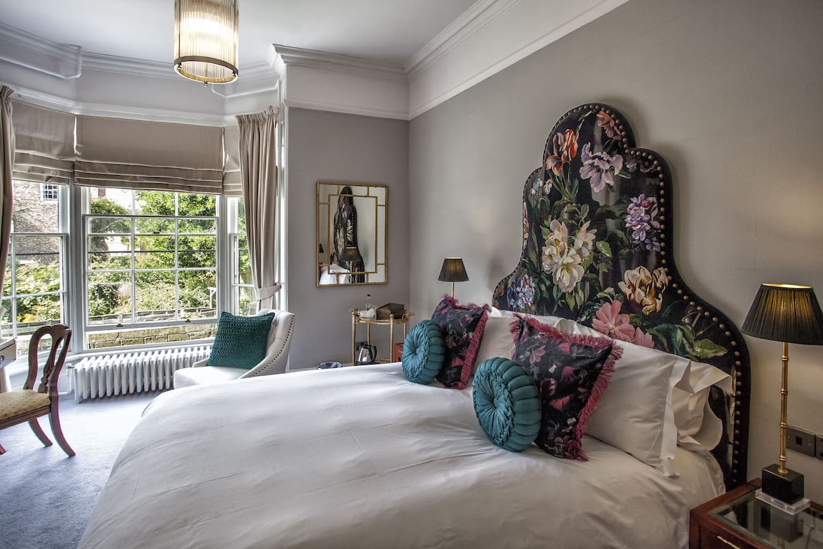A bedroom with a large bed, a nightstand, a chair, a window with curtains, and a floral headboard.