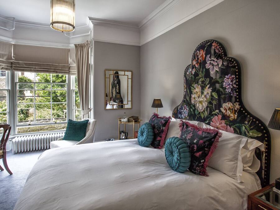 Een slaapkamer met een groot bed, een nachtkastje, een stoel, een raam met gordijnen en een hoofdbord met bloemenprint.