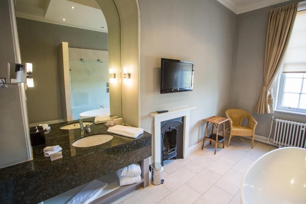 A modern bathroom with a large mirror, dual sinks, a flat-screen TV, a fireplace, a wicker chair, and a bathtub.