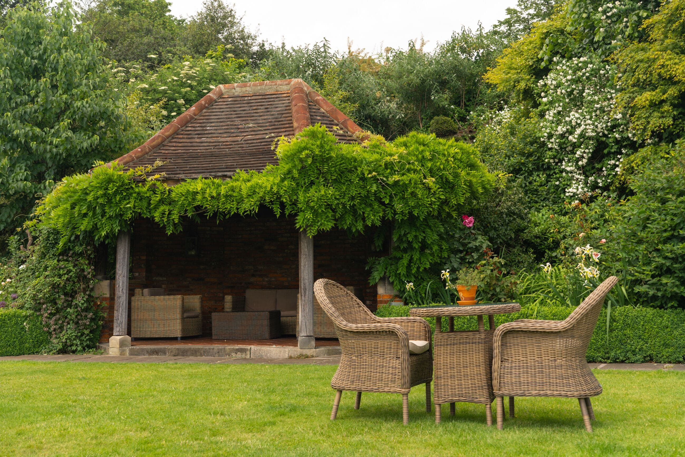 Een tuin met een prieel, rieten meubilair en weelderige begroeiing.