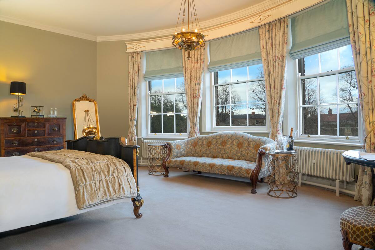 A bedroom with a large bed, a sofa, a small table, and a mirror. There are large windows with curtains and a chandelier.