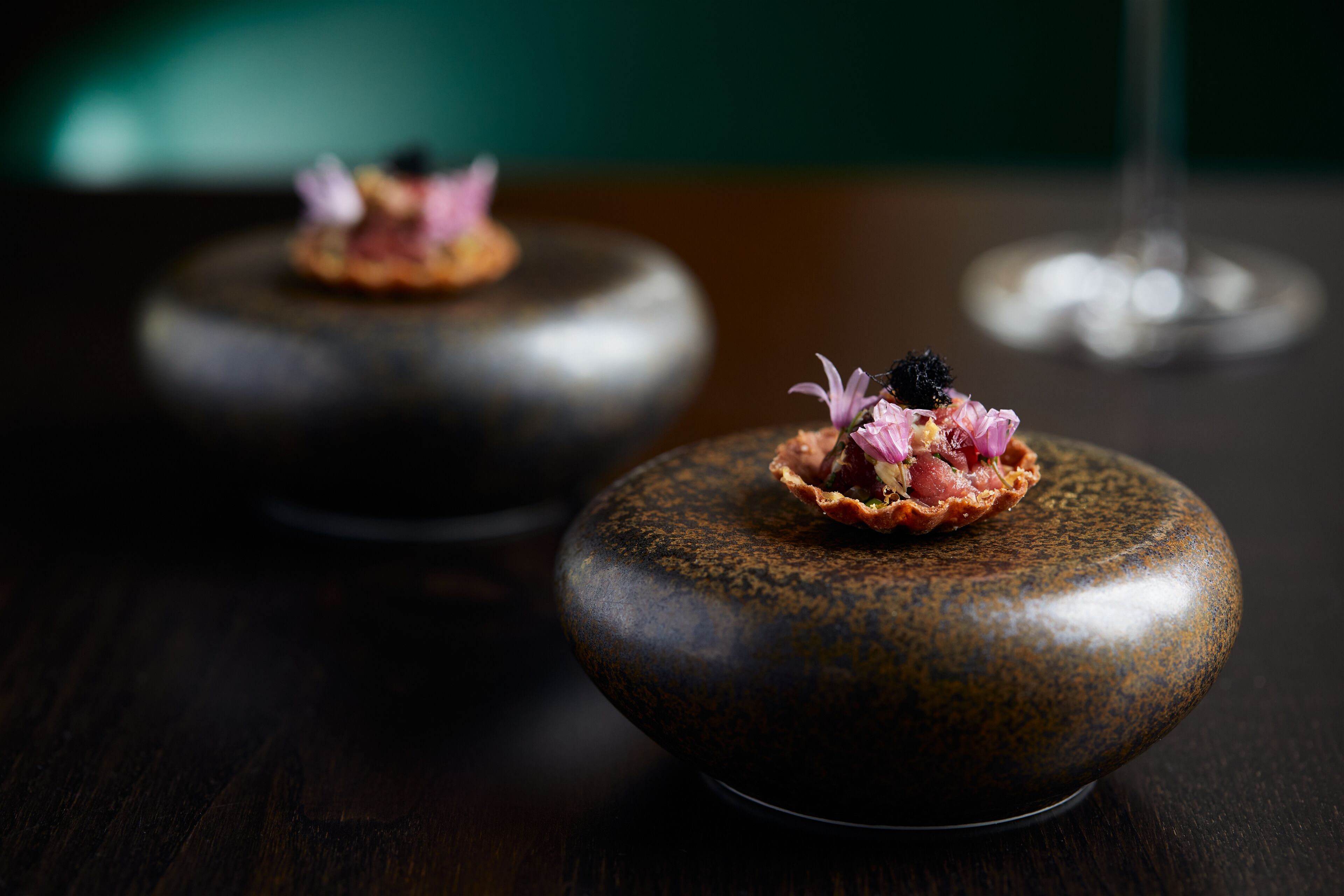 A table with two round, dark-colored ceramic dishes, each topped with a small tart and pink flowers.