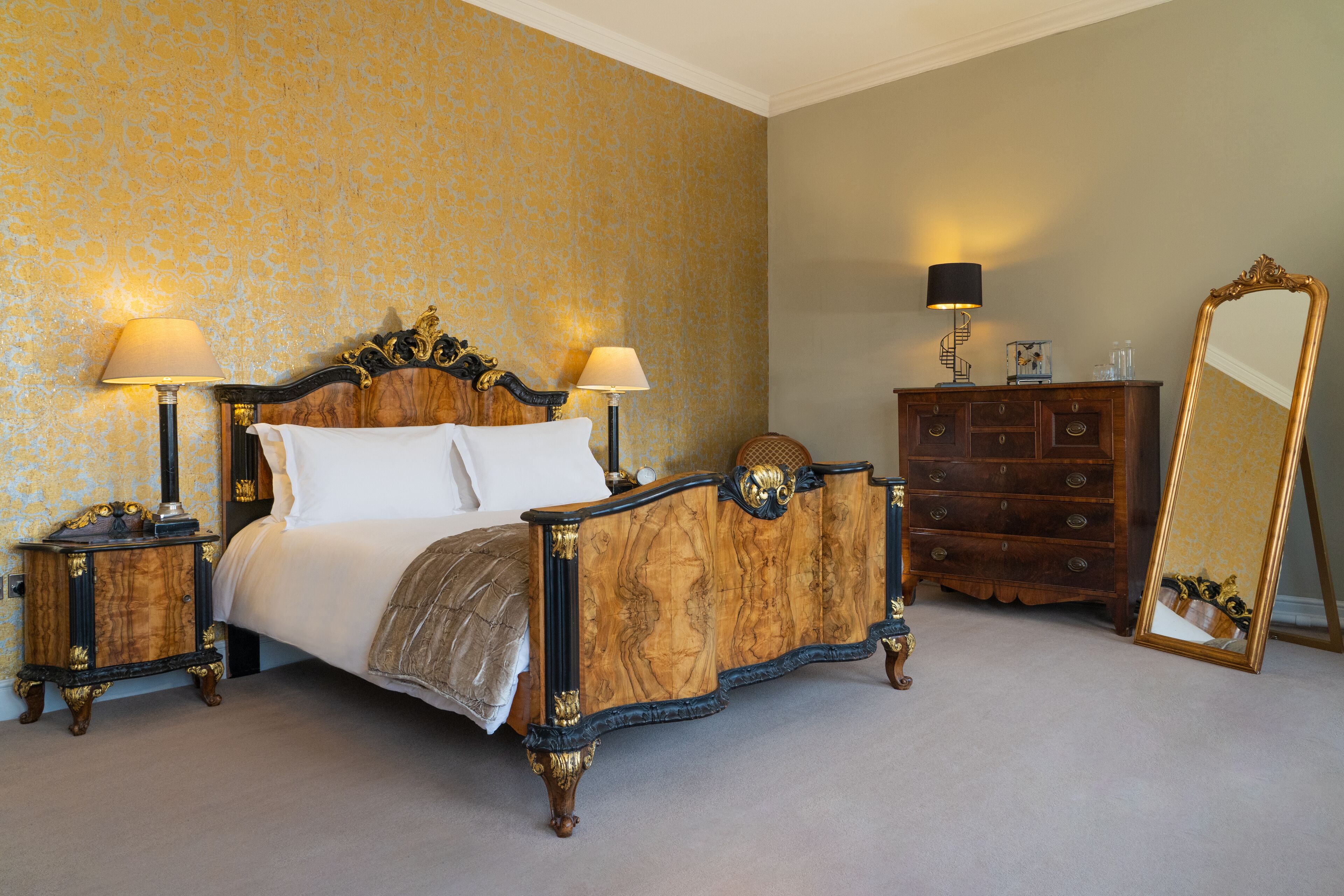 A classic four-poster bed with ornate woodwork and two bedside lamps, a wooden dresser with a mirror, and a patterned wallpaper in a hotel room.