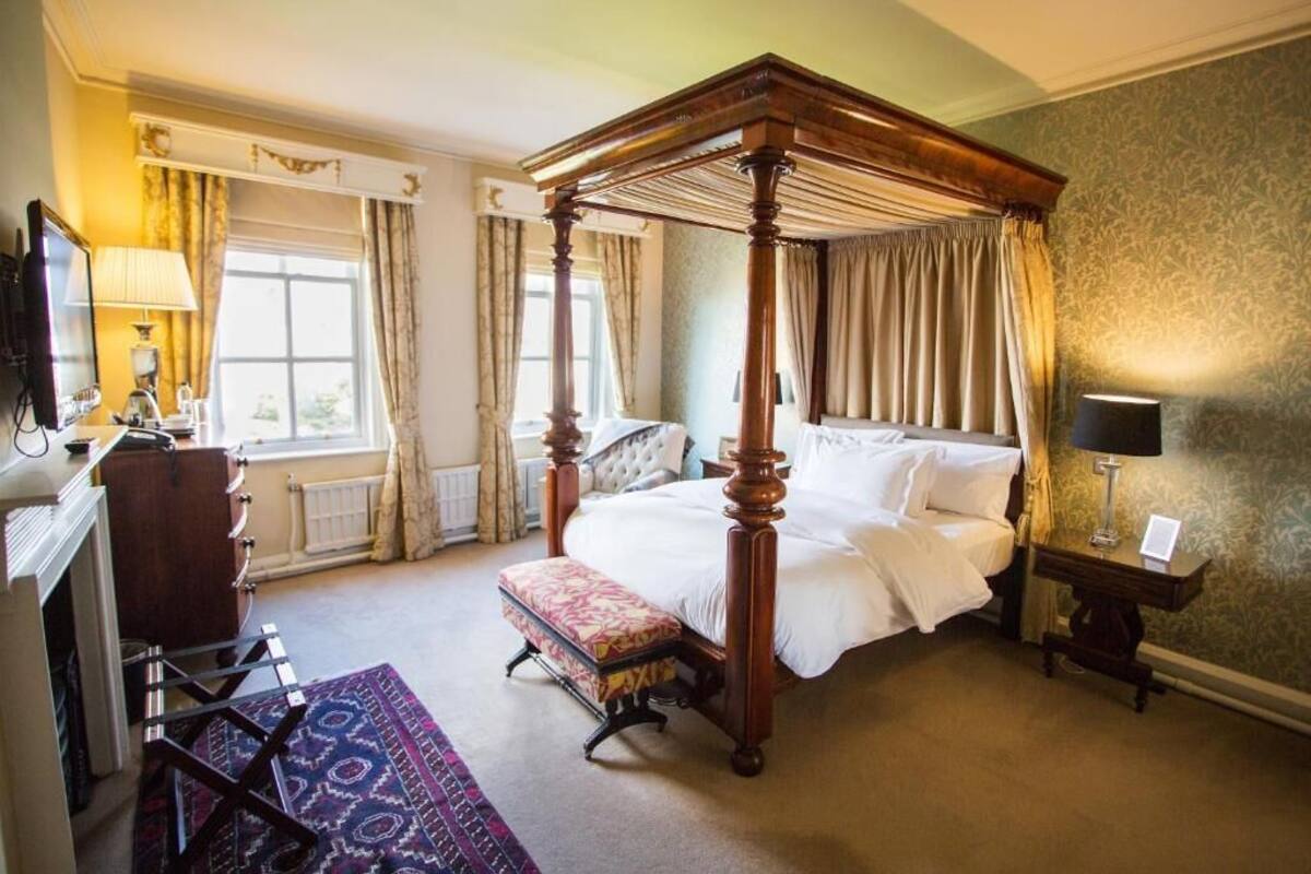 A four-poster bed with a matching canopy, a desk with a lamp, a chair, and a patterned rug in a hotel room.