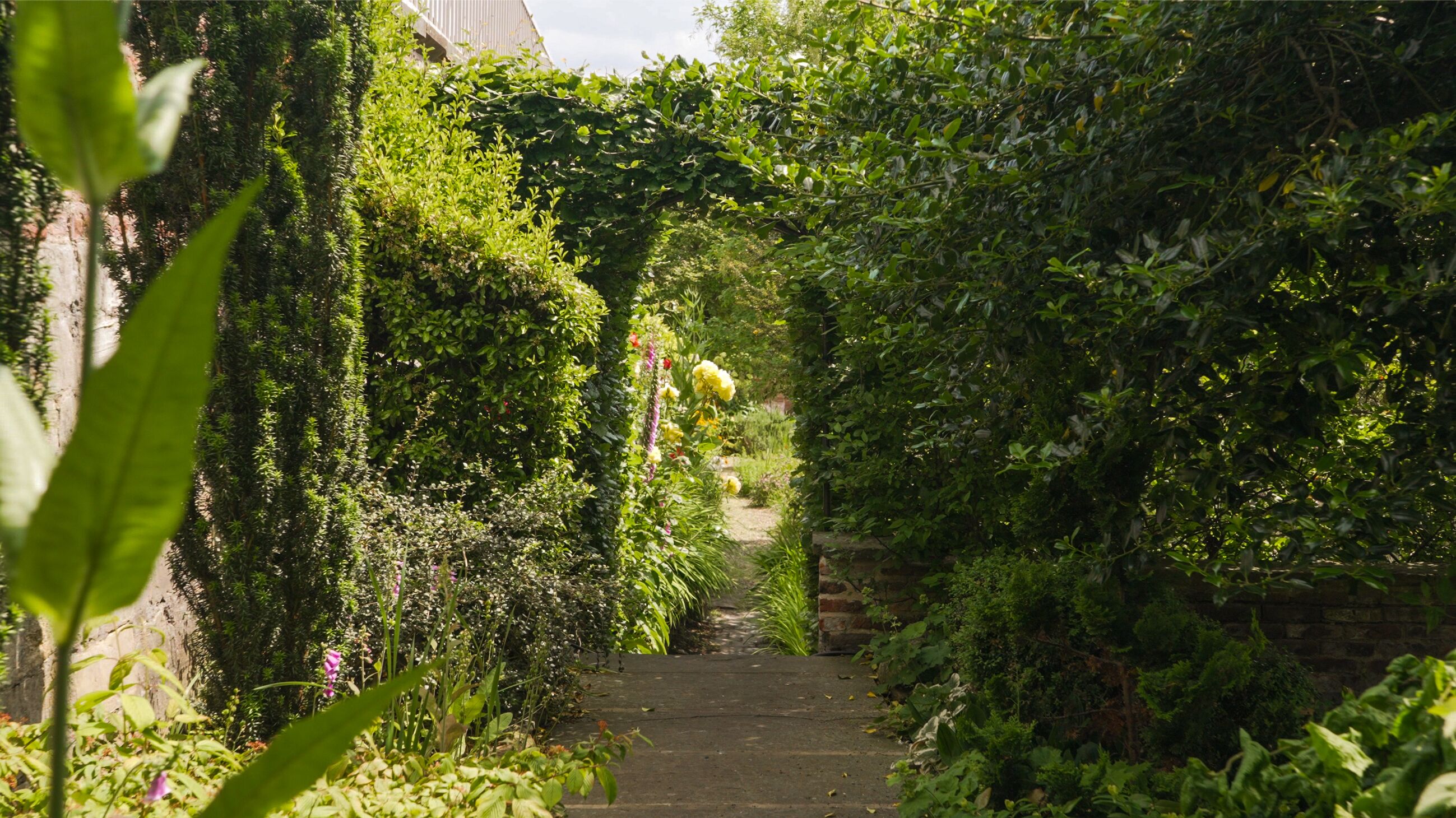 Un étroit sentier de pierre, bordé d’une végétation luxuriante et de plantes en fleurs, mène à l’entrée d’un bâtiment.