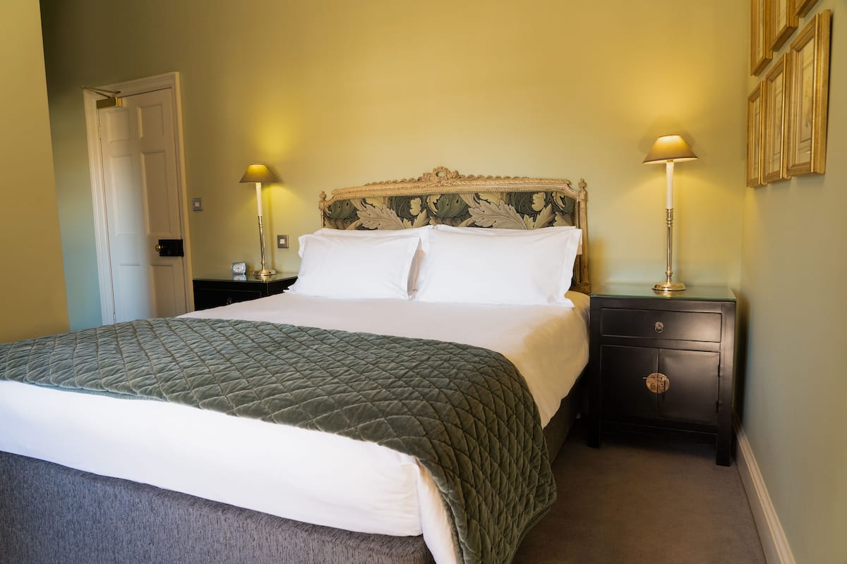 A neatly made bed with a quilted green blanket, a wooden headboard with leaf patterns, two bedside lamps, and a dark wooden nightstand with a drawer.