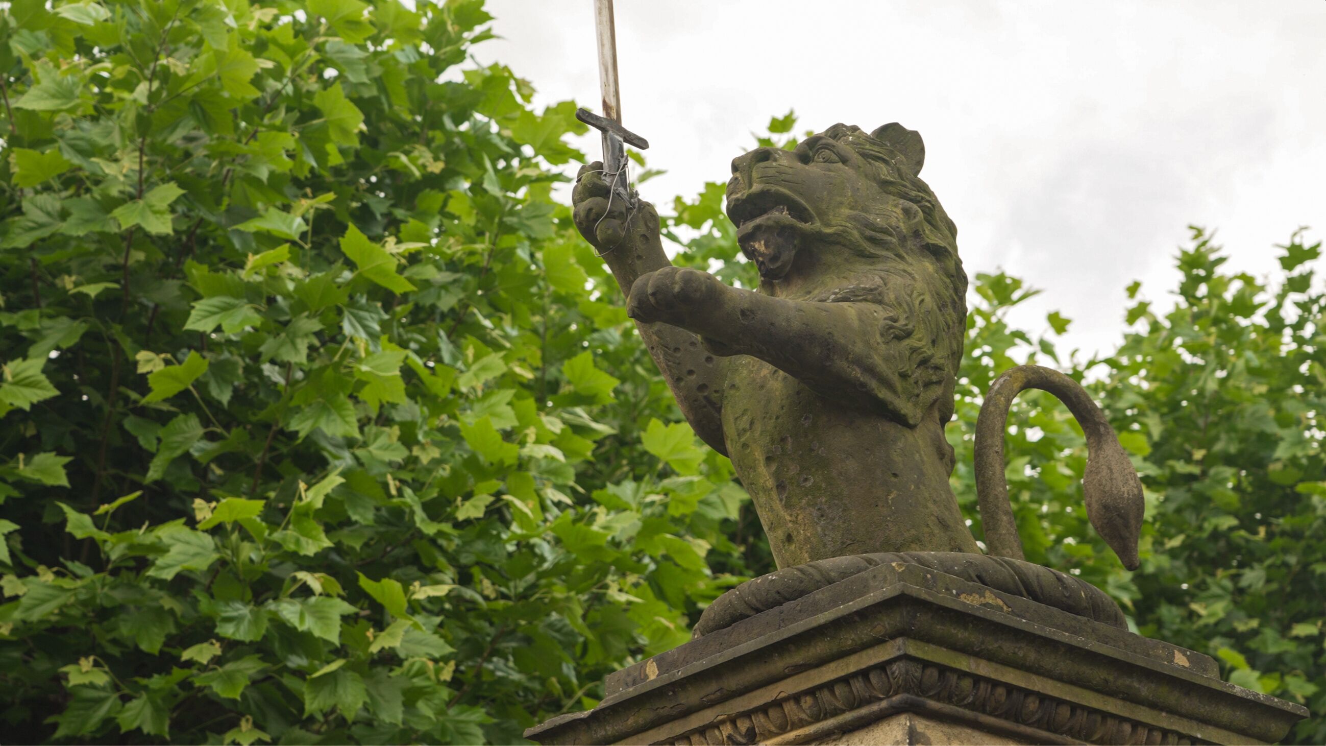 A lion statue holding a cross, set on a pedestal with a stone base, surrounded by green foliage.