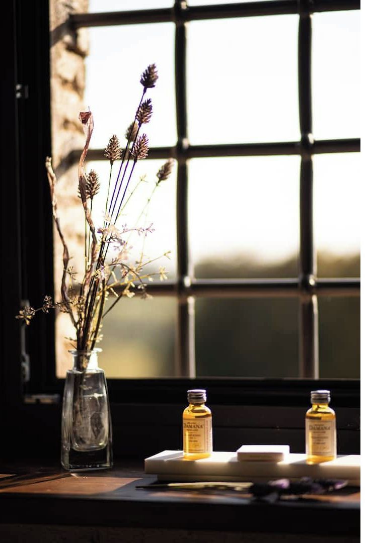 Une fenêtre donnant sur un immeuble, un vase contenant des fleurs séchées et deux bouteilles posées sur une table.