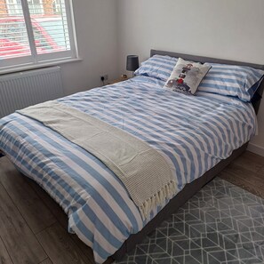 A bedroom with a bed, a bedside table, a lamp, and a window with blinds. - Captivating 2-bed Apartment in Bridlington (Bridlington)