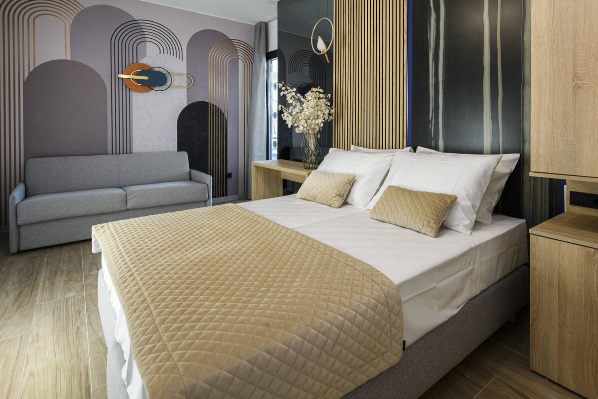 Un dormitorio moderno con una cama grande, un sofá gris y una mesita de noche de madera con un jarrón de flores.