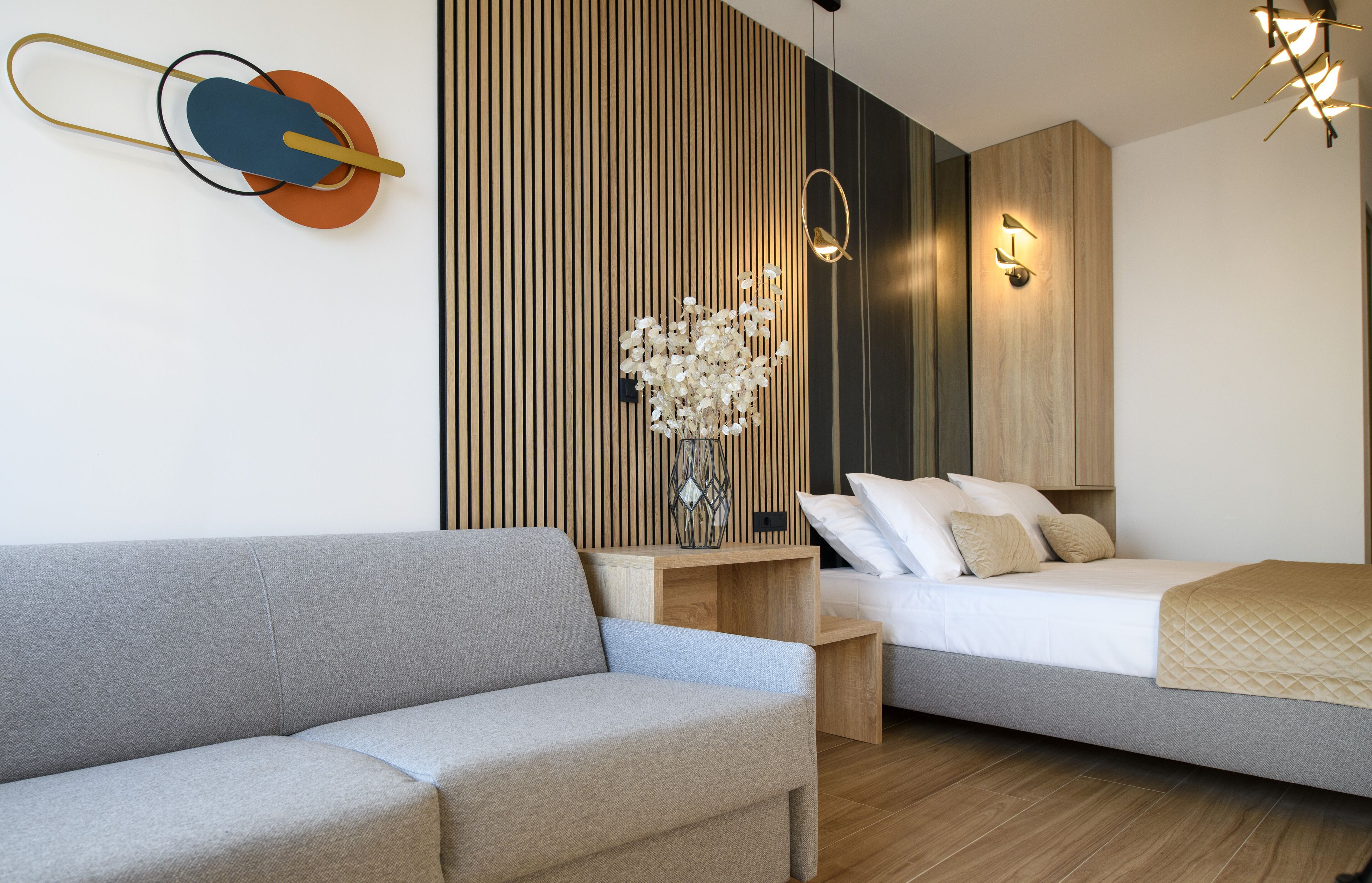 Un hotel moderno con un sofá gris, una mesita de noche de madera con un jarrón de flores, una cama con ropa blanca y una lámpara de pared.