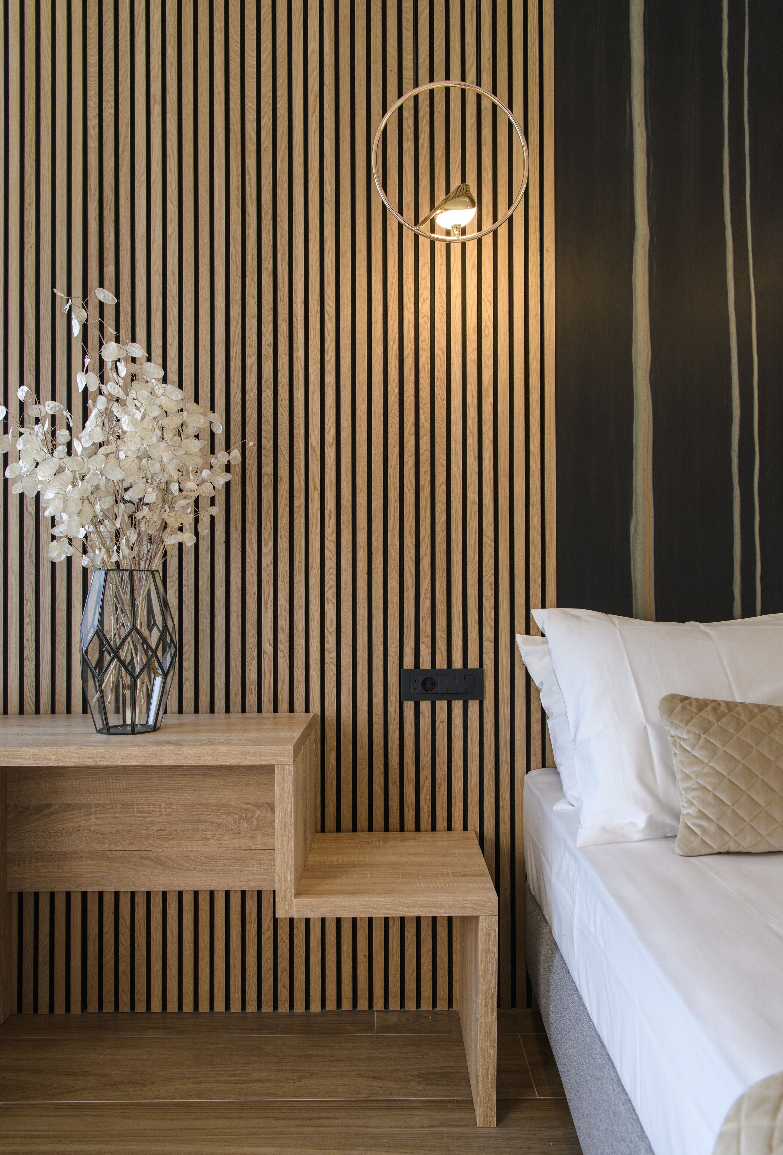 A bedroom with a wooden slat wall, a bedside table with a vase of flowers, a bed with white bedding, and a wall-mounted light fixture.