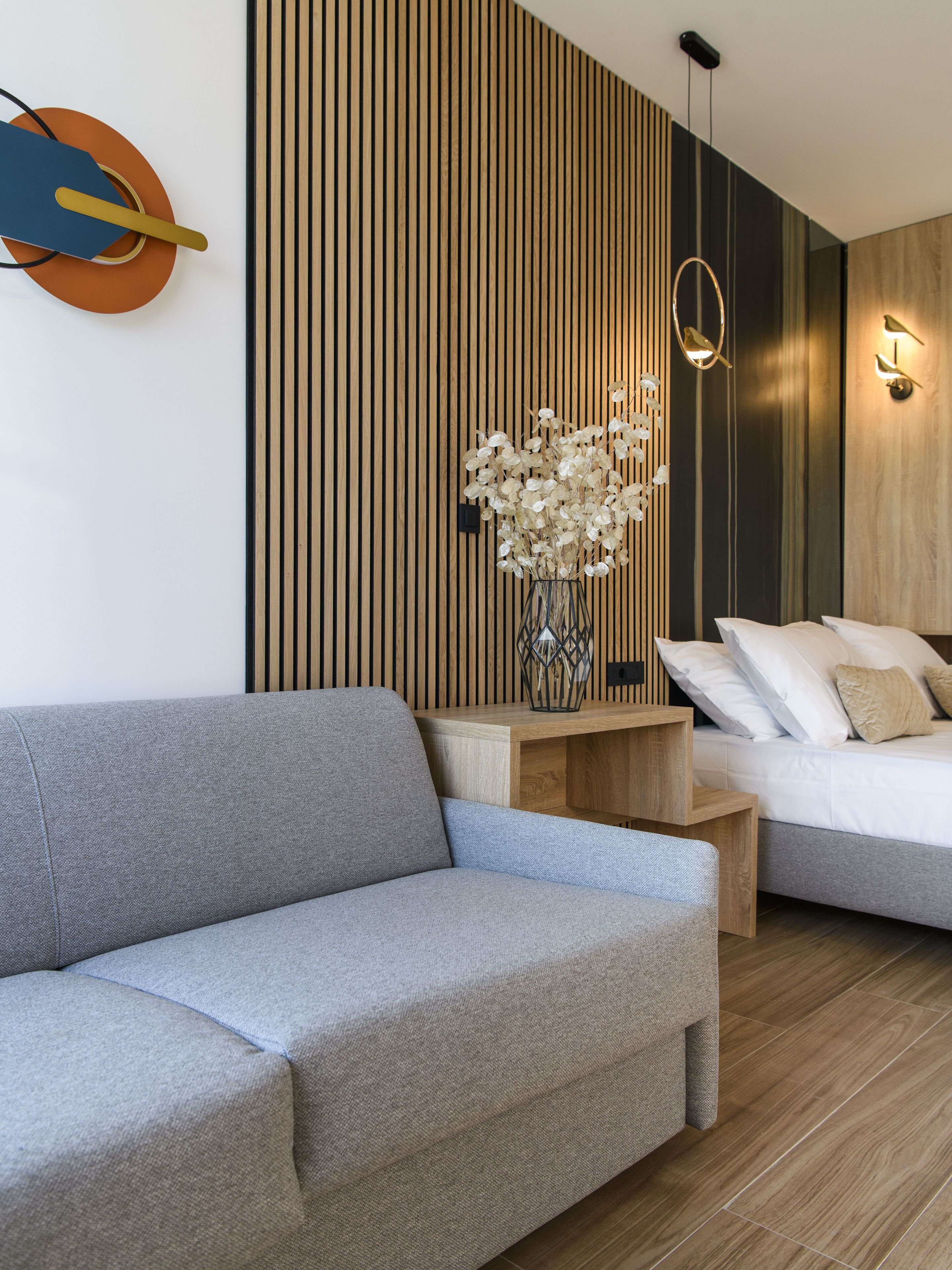 A modern bedroom with a wooden slat wall, a grey sofa, a wooden bedside table, a bed with white bedding, and a vase with dried flowers.