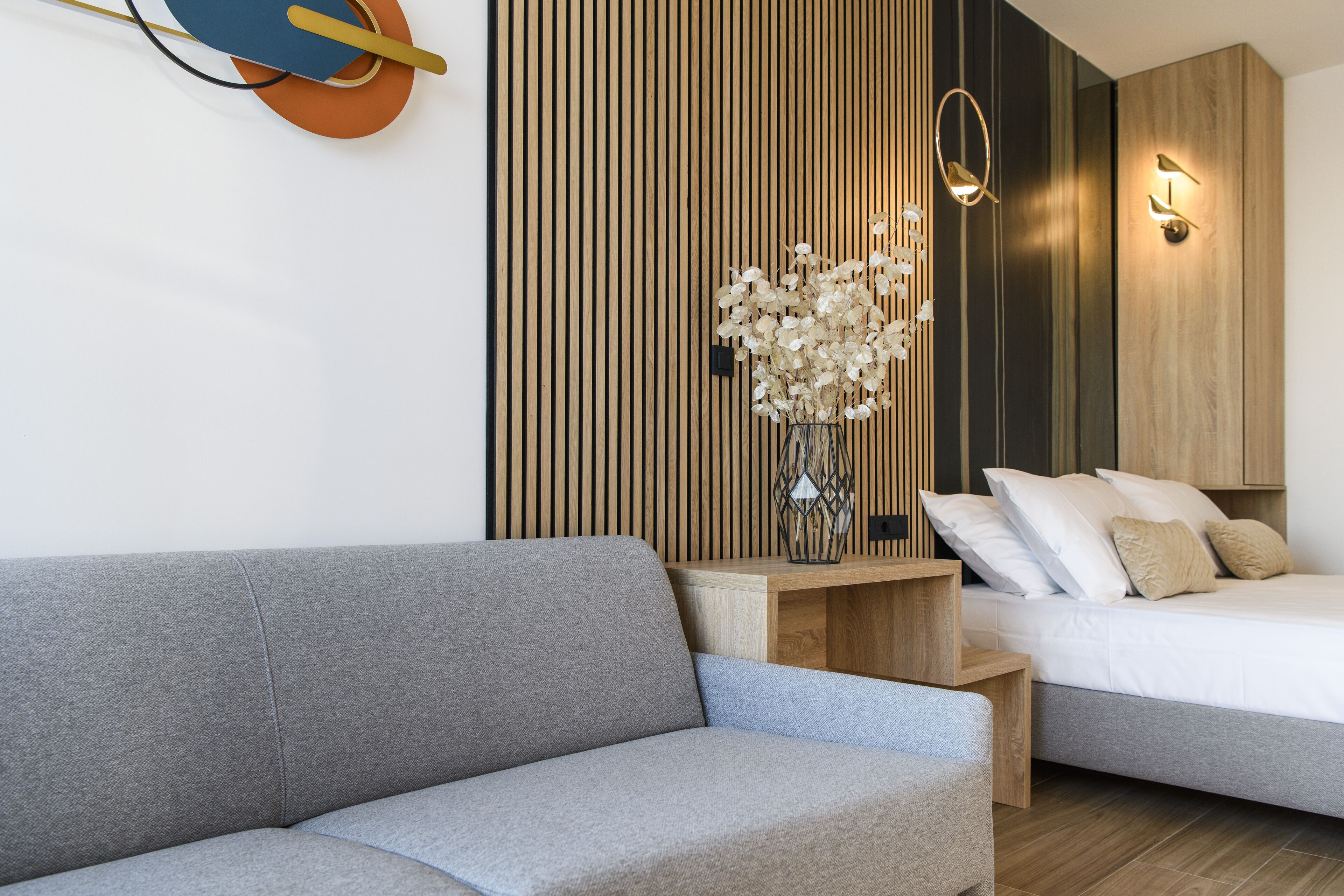 A modern bedroom with a grey sofa, a wooden bedside table with a vase of flowers, a bed with white and beige pillows, and a wall-mounted light fixture.