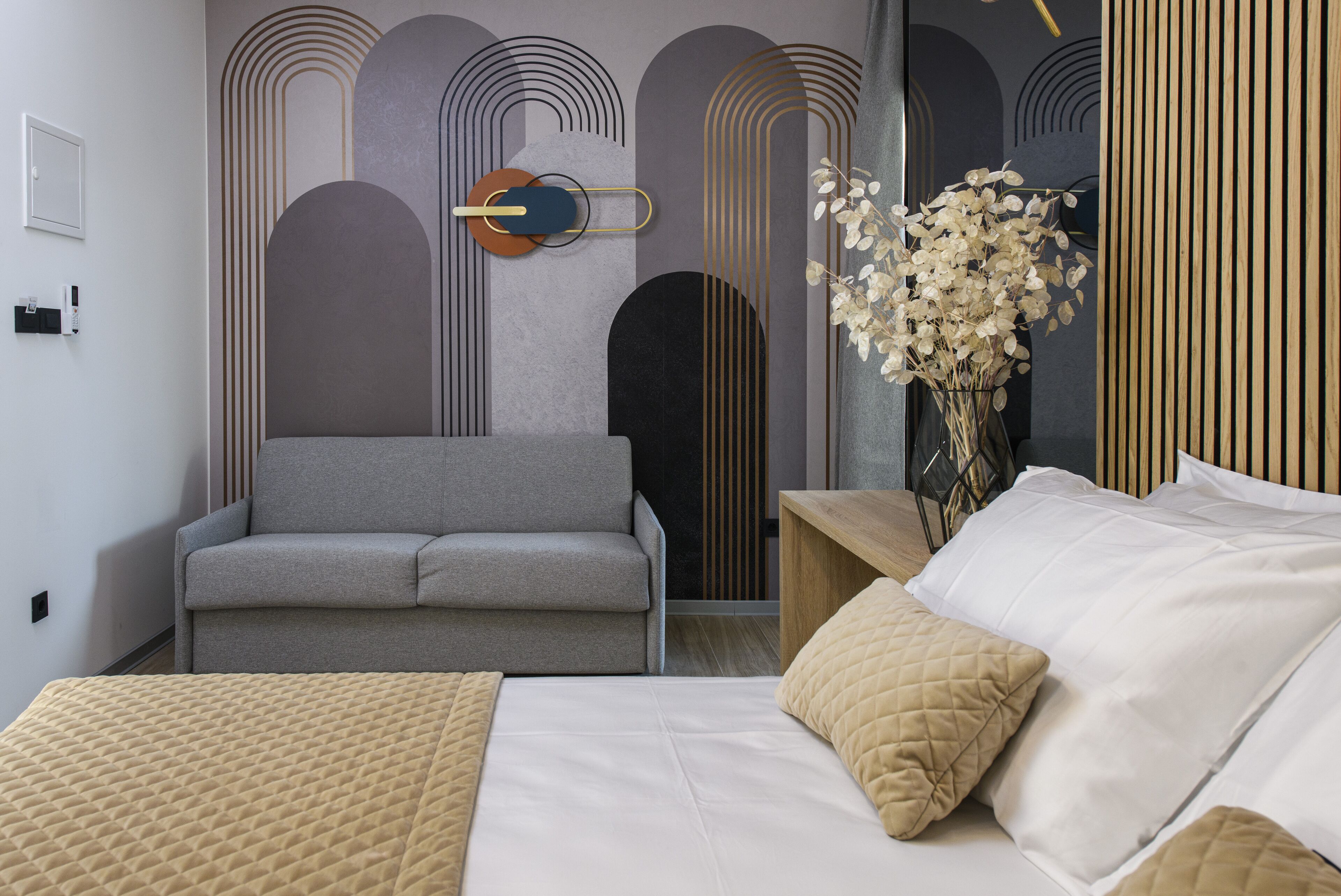 Una habitación de hotel moderna con un sofá gris, una mesita de madera con un jarrón de flores blancas, y una cama con ropa de cama blanca.