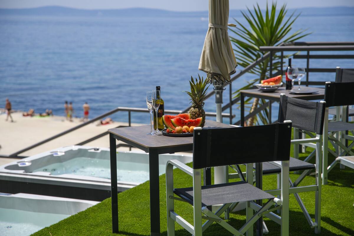 Mesa junto a la piscina con frutero, botella de vino y copa, con vista a una playa y al océano.