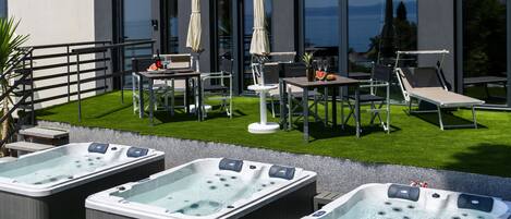 A modern outdoor patio with three hot tubs, a dining area with chairs and tables, and a view of the sea through large windows.