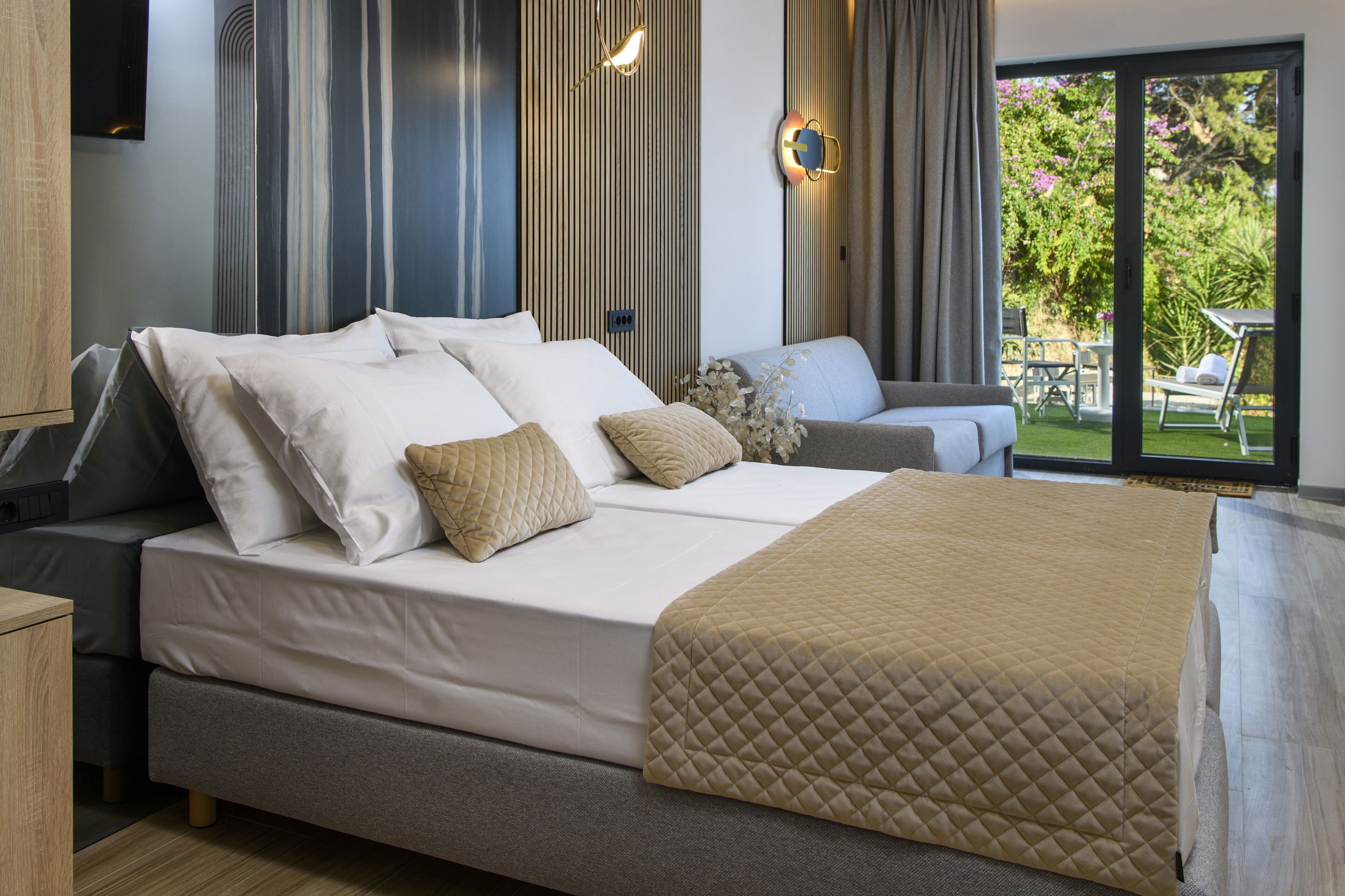 A modern hotel room with a large bed, a sofa, and a view of the garden through the sliding glass door.