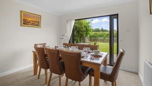 A dining area with a table set for six, six brown chairs, a painting on the wall, and a view of a garden through a sliding glass door. - House - Sleeps 6 & Free Parking, Pets (Great Yarmouth)